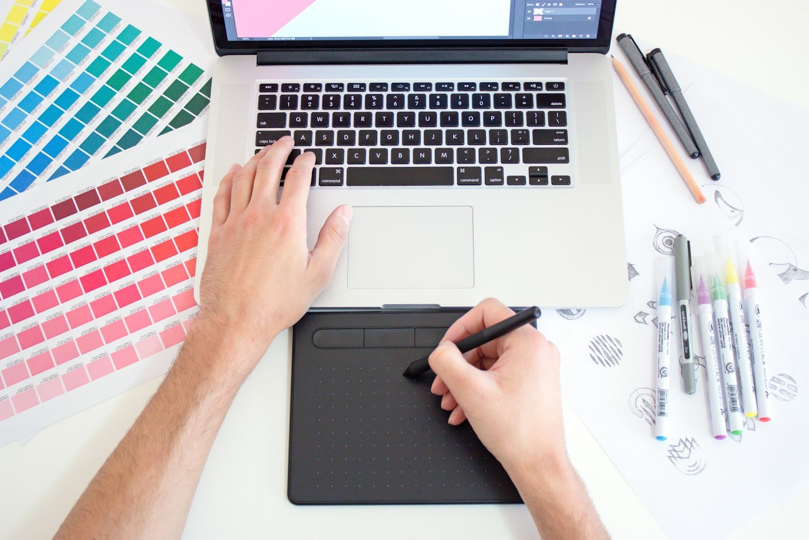 A designer's workspace shot from above: laptop, drawing tablet, colour palette swatches and pens laid out in natural daylight, hands sketching mid-project